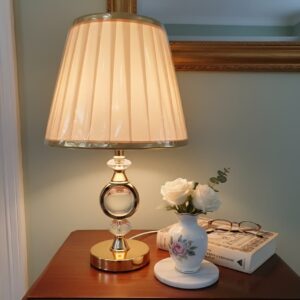 Lámpara de mesa moderna con base de cristal y metal, y pantalla de tela plisada.